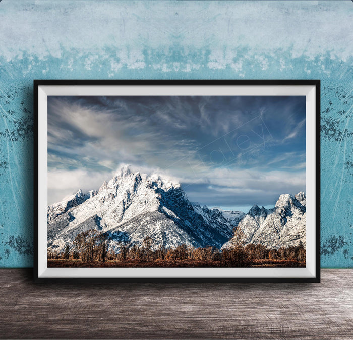 A framed photograph of a snow-capped mountain range against a cloudy sky, displayed on a wooden surface against a blue textured wall.