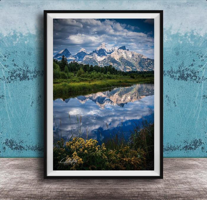 A framed photograph of a serene landscape with a mountain range reflected in a calm lake, surrounded by lush greenery and vibrant yellow flowers.