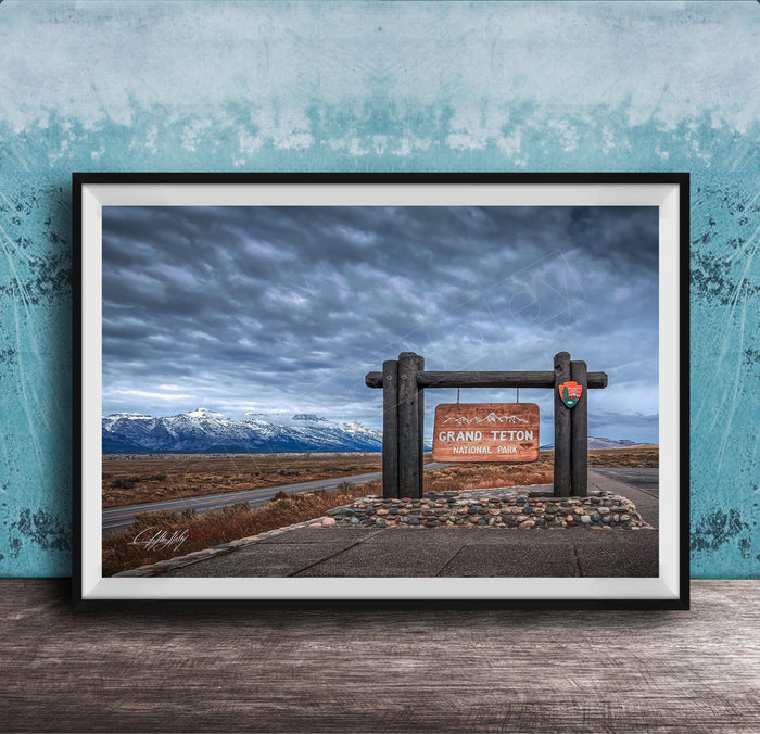 A framed photograph of a sign for Grand Teton National Park, with a cloudy sky and mountains in the background.