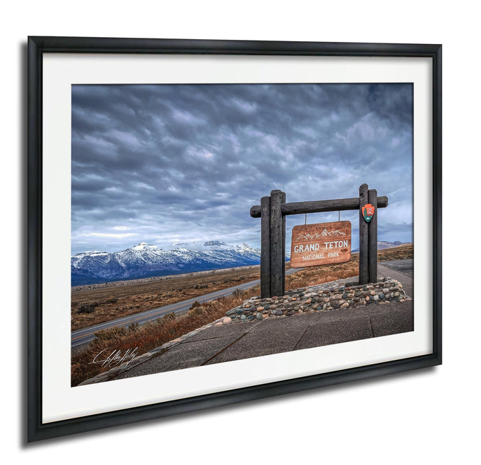 A framed photograph of a scenic landscape with a wooden sign reading "Grand Teton National Park" in the foreground, set against a backdrop of snow-capped mountains and a cloudy sky.