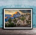The image is a framed photograph depicting a mountainous landscape with a large mountain peak in the background, surrounded by trees and rocky terrain. The sky is filled with warm, golden light, creating a picturesque and serene atmosphere.