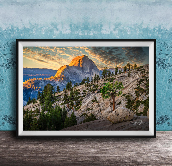 The image is a framed photograph depicting a mountainous landscape with a large mountain peak in the background, surrounded by trees and rocky terrain. The sky is filled with warm, golden light, creating a picturesque and serene atmosphere.