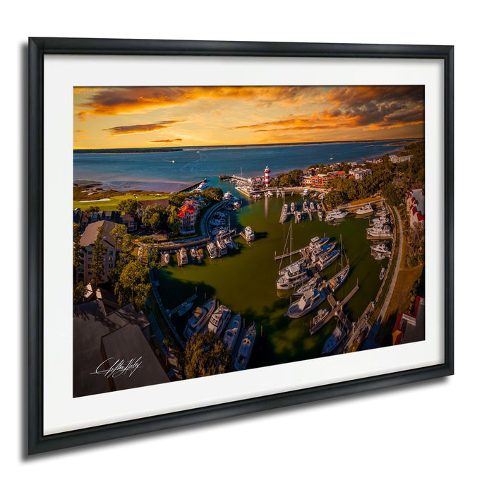 The image is a framed aerial view of a harbor with boats docked at the marina, set against a backdrop of a colorful sunset sky.