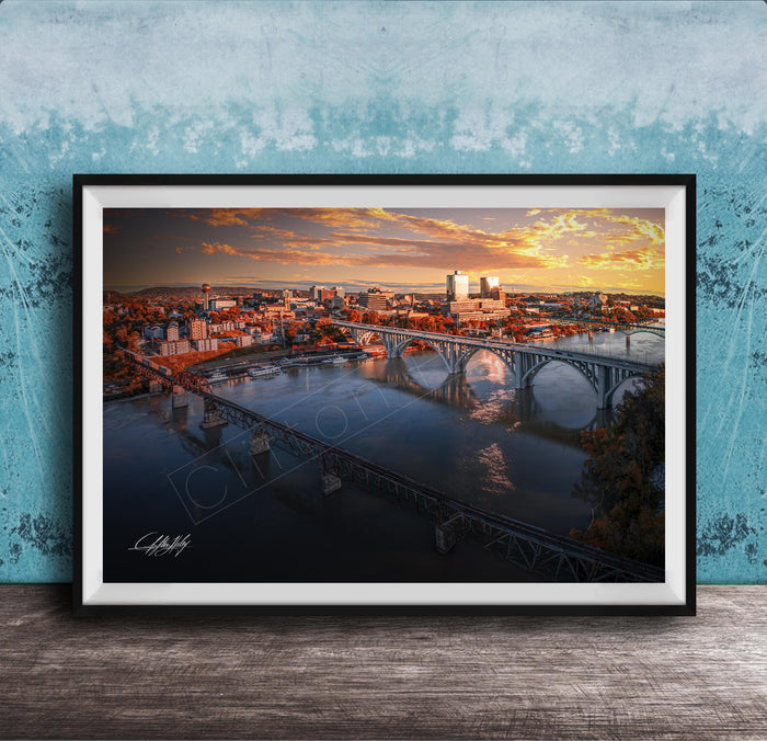 The image is a framed photograph depicting a cityscape with a bridge spanning a river, set against a backdrop of a colorful sunset sky.