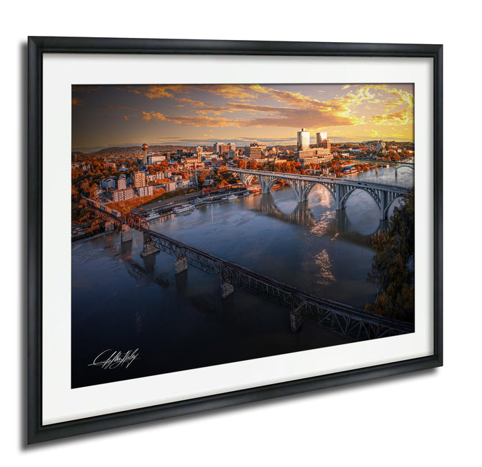 The image is a framed aerial view of a cityscape with a bridge spanning a river. The bridge is surrounded by buildings and trees, and the sky is filled with clouds, creating a dramatic backdrop. The photograph is framed with a black border and the artist's signature is visible in the bottom left corner.