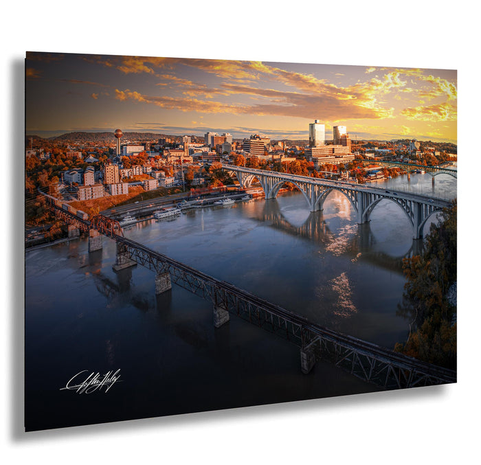 a cityscape with a large bridge spanning across a river, reflecting the warm hues of the setting sun. The sky is filled with vibrant colors, creating a picturesque scene.