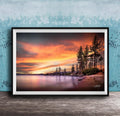 A framed photograph depicting a serene landscape with a calm lake, a rocky shoreline, and a line of trees silhouetted against a vibrant sunset sky.