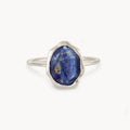 A sterling silver lapis lazuli mood ring by bluboho sits centered against a white background, featuring a raw, oval-shaped blue gemstone adorned with unique golden specks. The gemstone's irregular surface enhances its rustic charm, while the simple and delicate band highlights its beauty.