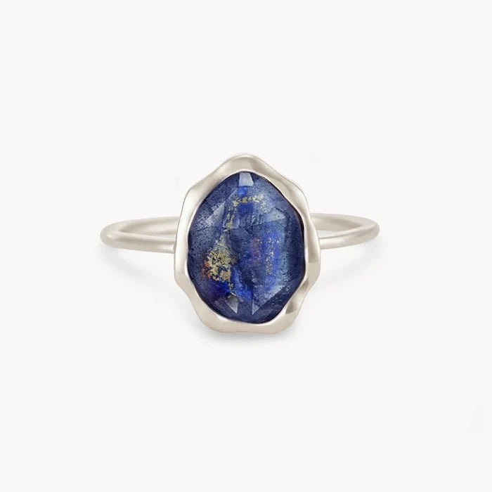 A sterling silver lapis lazuli mood ring by bluboho sits centered against a white background, featuring a raw, oval-shaped blue gemstone adorned with unique golden specks. The gemstone's irregular surface enhances its rustic charm, while the simple and delicate band highlights its beauty.