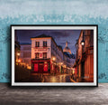 a charming street scene in Paris, France, with a row of colorful buildings lining the cobblestone street. The buildings are illuminated by warm street lamps, creating a cozy and inviting atmosphere. The sky above is a deep blue, providing a beautiful backdrop to the scene. The image is framed and displayed on a wooden surface, adding a rustic touch to the overall composition.