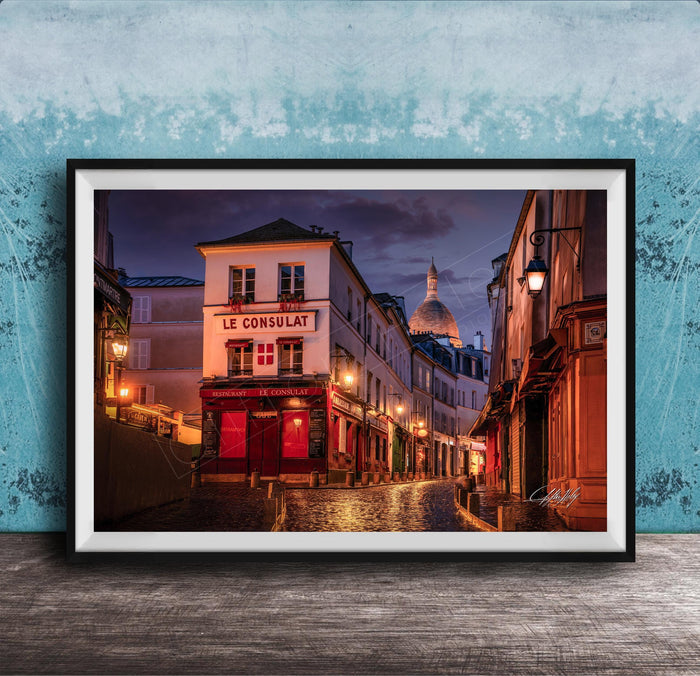 a charming street scene in Paris, France, with a row of colorful buildings lining the cobblestone street. The buildings are illuminated by warm street lamps, creating a cozy and inviting atmosphere. The sky above is a deep blue, providing a beautiful backdrop to the scene. The image is framed and displayed on a wooden surface, adding a rustic touch to the overall composition.