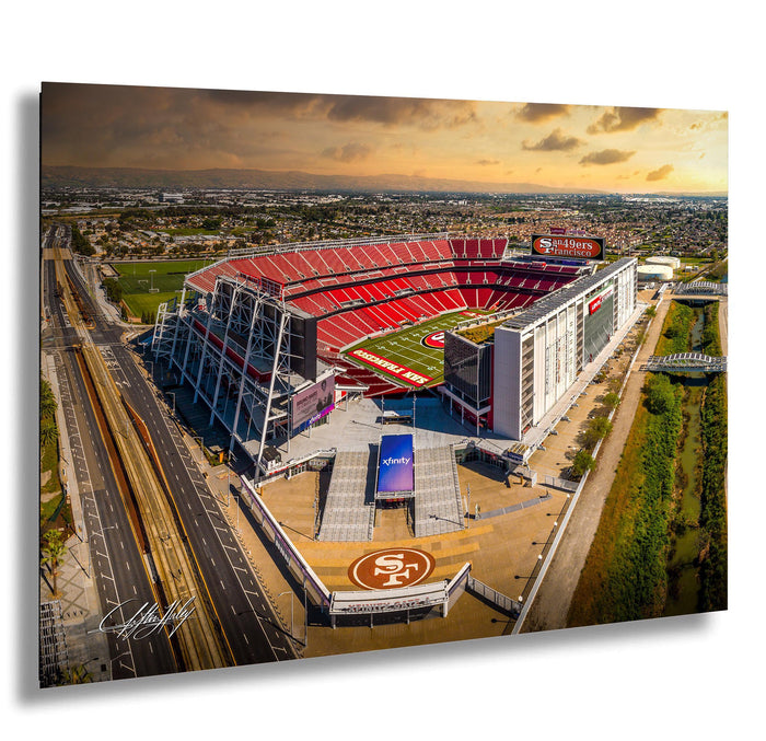 An aerial view of a large sports stadium, likely the San Francisco 49ers stadium, with the surrounding cityscape and a cloudy sky in the background.