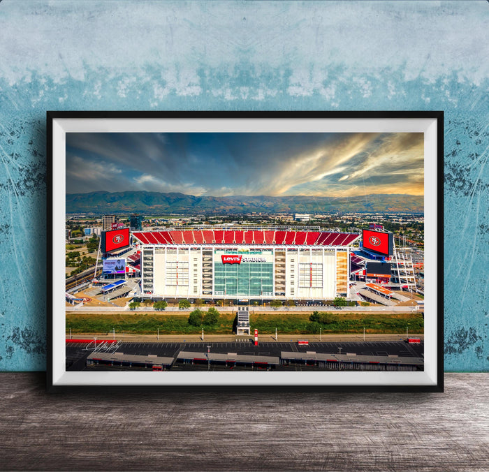 A framed photograph of a large sports stadium with a red roof, surrounded by mountains and a cloudy sky.