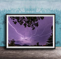 A framed photograph depicting a dramatic lightning storm with vibrant purple and blue hues illuminating the night sky, framed against a blue wall.
