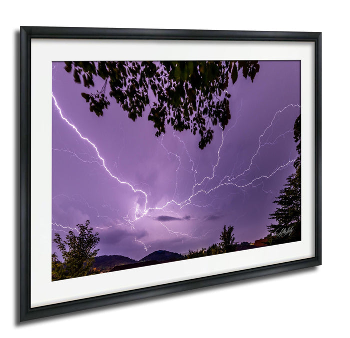 A framed photograph depicting a dramatic lightning storm with bright bolts of light illuminating the sky and silhouetting trees in the foreground.
