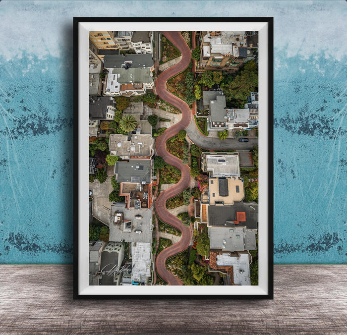 The image is an aerial view of a winding road that curves through a cityscape, with buildings and greenery lining the sides of the road.