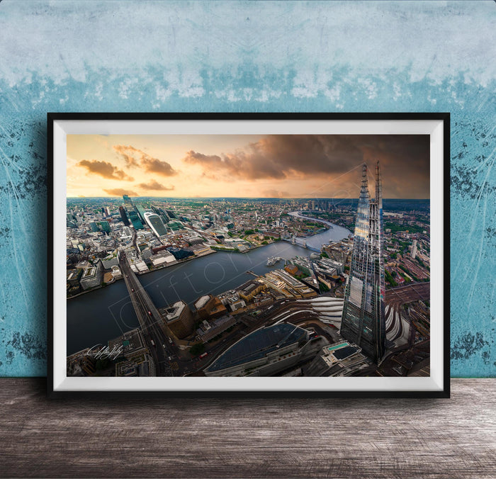 The image is a framed aerial view of a cityscape, showcasing a mix of tall buildings, roads, and a river. The sky is filled with clouds, creating a dramatic backdrop for the city's skyline. The image is framed and displayed on a wooden surface, likely a wall or shelf.