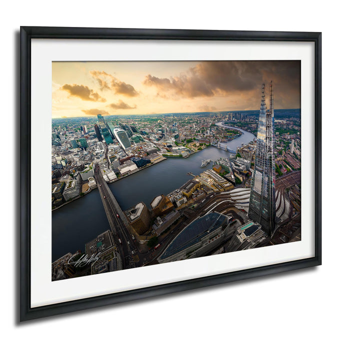 The image is a framed aerial view of a cityscape, featuring a mix of tall buildings, roads, and a river. The sky is filled with clouds, creating a dramatic backdrop for the city's skyline. The image is framed in a black frame and is hung on a wall.