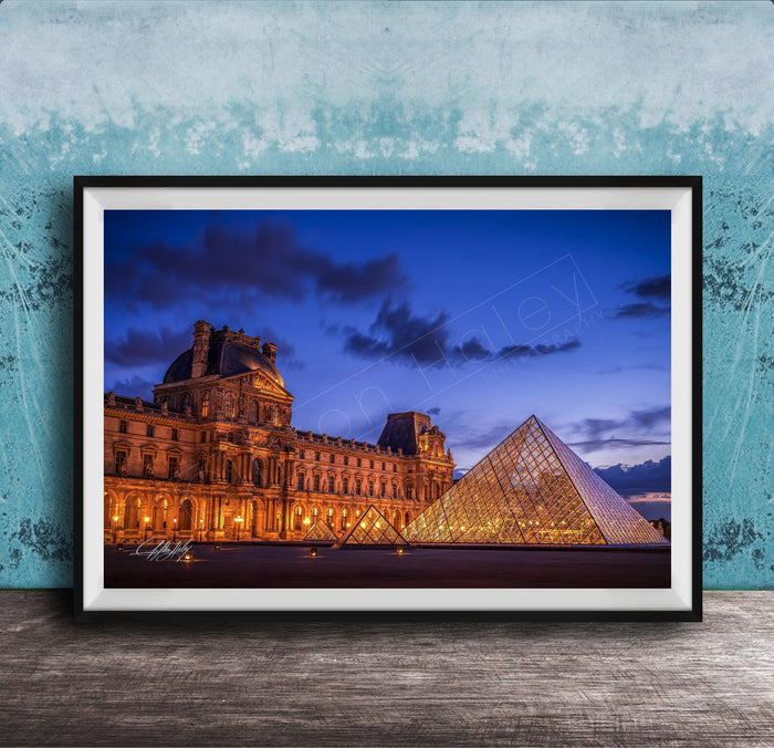 the Louvre Museum in Paris, France, with its iconic glass pyramid structure illuminated at night against a dark blue sky.