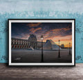A framed photograph of the Louvre Museum in Paris, France, with the iconic glass pyramid structure of the Louvre Museum prominently featured against a dramatic sky at sunset.