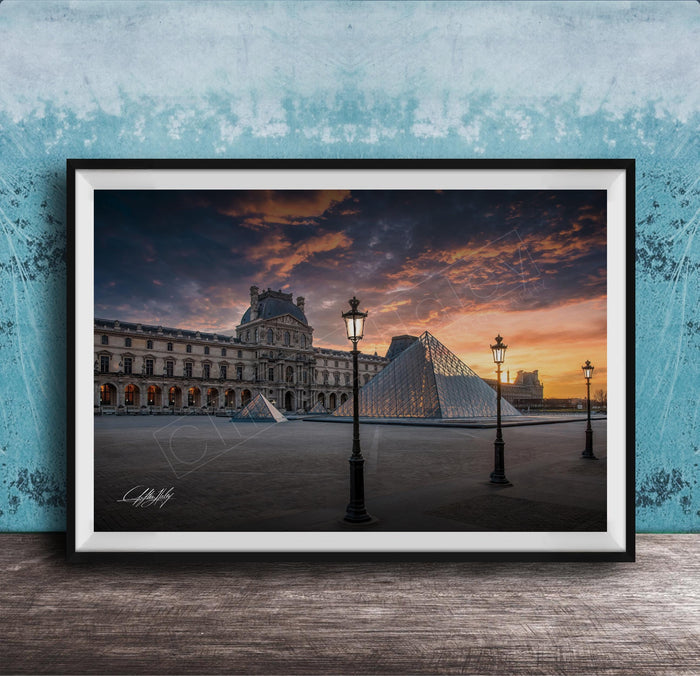 A framed photograph of the Louvre Museum in Paris, France, with the iconic glass pyramid structure of the Louvre Museum prominently featured against a dramatic sky at sunset.