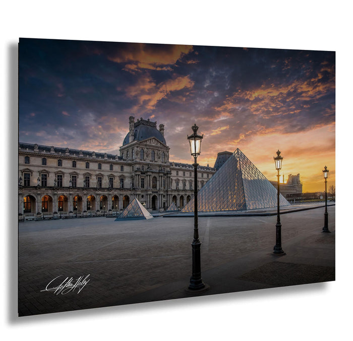 the Louvre Museum in Paris, France, with its iconic glass pyramid and surrounding architecture illuminated against a vibrant sunset sky.