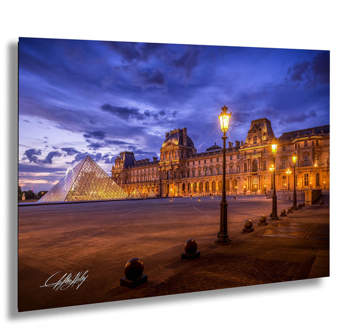 the Louvre Museum in Paris, France, with its iconic glass pyramid and illuminated architecture against a dark blue sky.