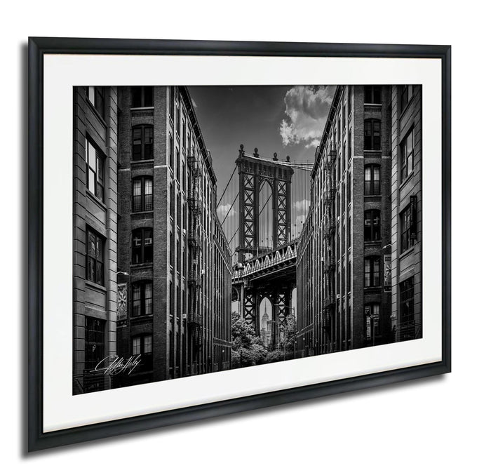 A black and white photograph of the Manhattan Bridge in New York City, with the bridge's intricate steel truss structure spanning across the frame between two tall buildings.