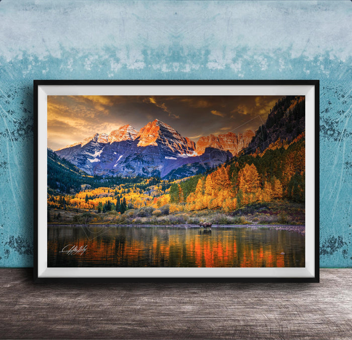 The image is a framed photograph depicting a scenic mountain landscape with a lake in the foreground, surrounded by trees with autumn foliage.
