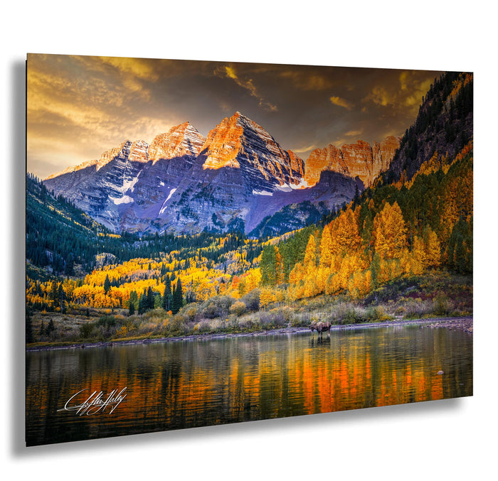 A breathtaking mountain landscape with a lake in the foreground, surrounded by vibrant fall foliage. The mountains are covered in snow and their peaks are bathed in warm, golden light, creating a stunning contrast against the cloudy sky. A moose can be seen in the distance, adding to the serene and picturesque scene.
