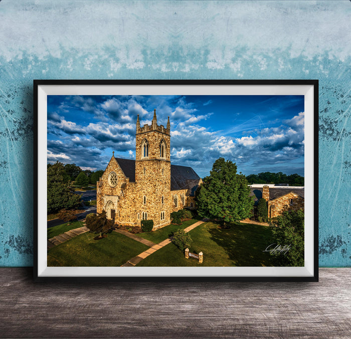 A stone church with a tall tower, surrounded by trees and a grassy area, set against a blue sky with clouds.