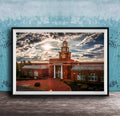 A framed photograph of a red brick building with a tower, set against a cloudy sky at sunset.