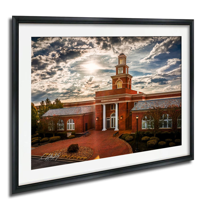 A framed photograph of a brick building with a prominent tower, set against a cloudy sky with the sun peeking through.