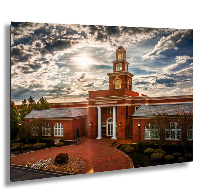 A large brick building with a prominent tower, surrounded by landscaping and set against a dramatic cloudy sky.