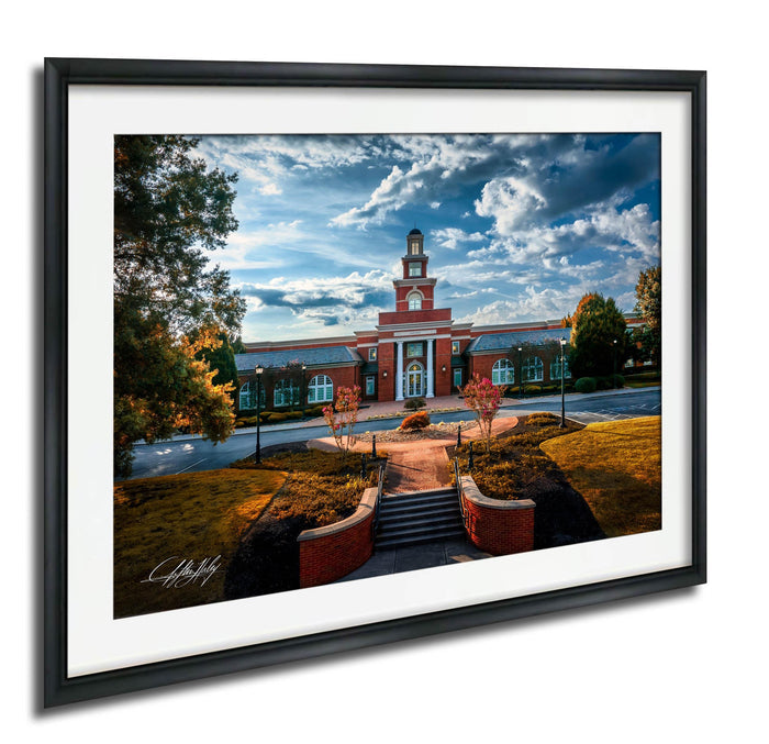 A framed photograph depicting a red brick building with a clock tower, surrounded by trees and a paved walkway leading up to it.