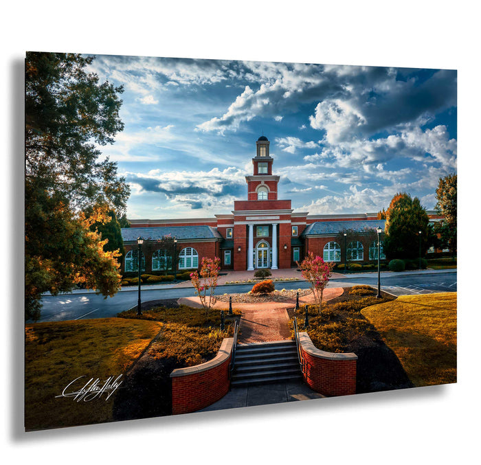A red brick building with a clock tower, surrounded by trees and a paved walkway leading up to it. The sky is blue with white clouds, and the scene is framed by trees with autumn foliage.