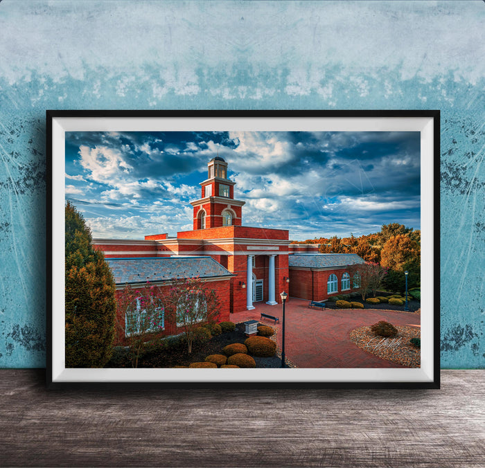 A framed photograph of a red brick building with a prominent tower, set against a blue sky with clouds.