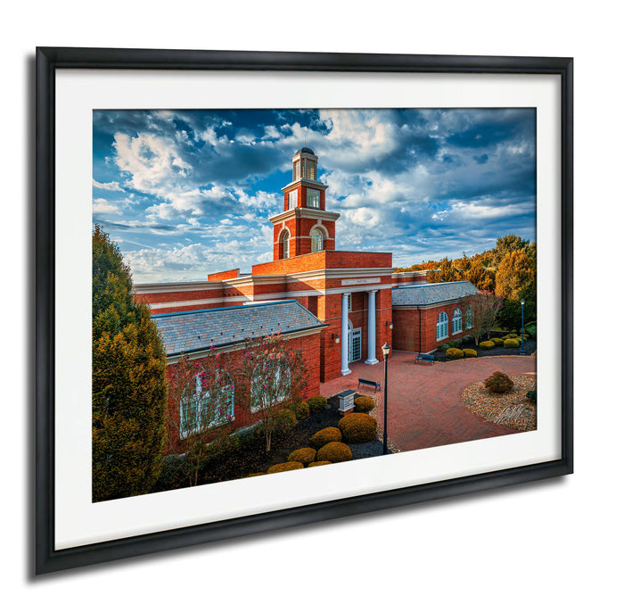 A framed photograph of a red brick building with a clock tower, surrounded by trees and landscaping, set against a blue sky with clouds.