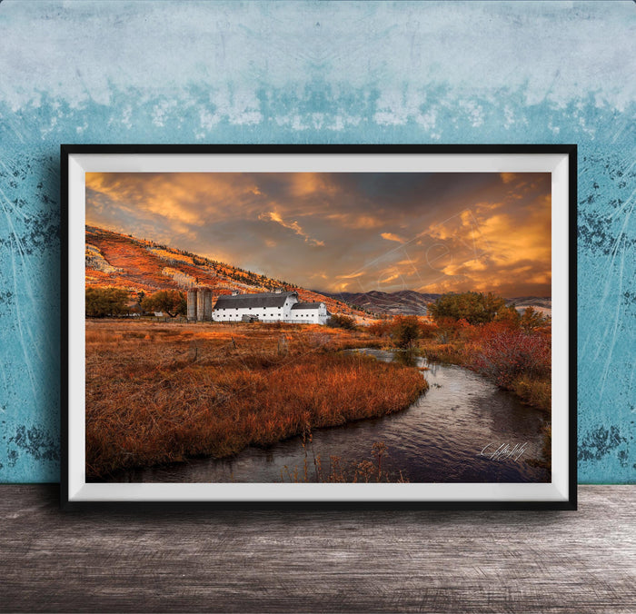 a picturesque rural landscape with a farmhouse nestled among rolling hills and a winding stream. The sky is filled with vibrant orange and yellow hues, creating a warm and serene atmosphere. The overall scene is a beautiful representation of nature and the tranquility of rural life.