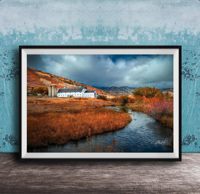 a picturesque rural landscape with a small white building nestled among the rolling hills and lush greenery. The scene is framed and displayed on a wooden surface, with a blue wall in the background.