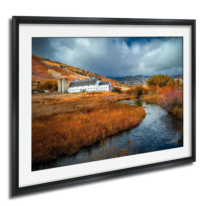 A framed photograph depicting a picturesque rural landscape with a farmhouse, a stream, and a hill covered in autumn foliage.
