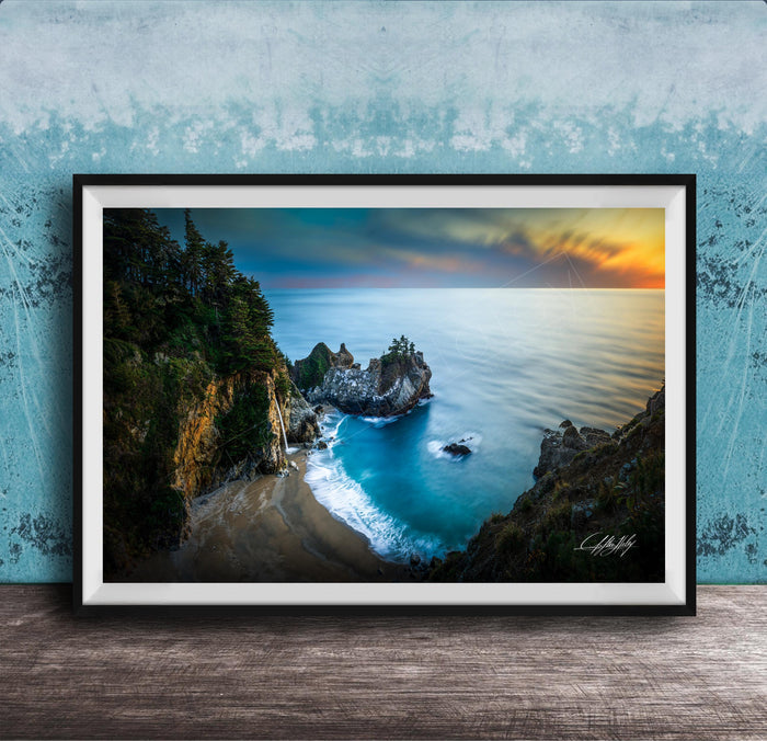 A framed photograph depicting a scenic coastal landscape with a rocky shoreline, a sandy beach, and a body of water with a sunset sky in the background.