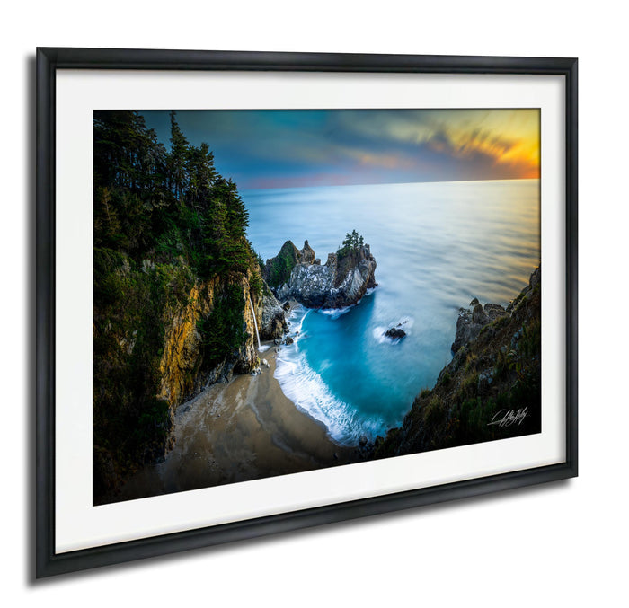 The image is a framed photograph depicting a scenic coastal landscape with a rocky cliff overlooking a beach and the ocean.