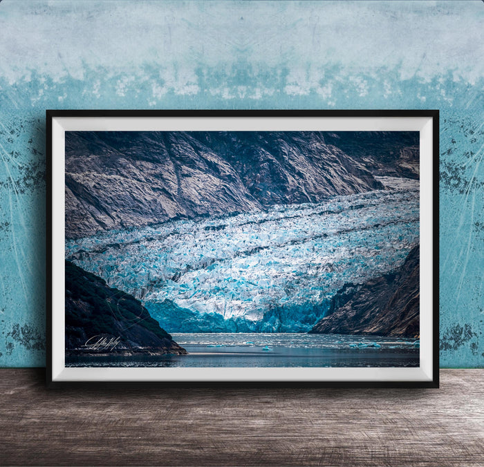 A framed photograph of a large, majestic glacier with a blue-green hue, set against a backdrop of rugged mountains and a serene body of water.