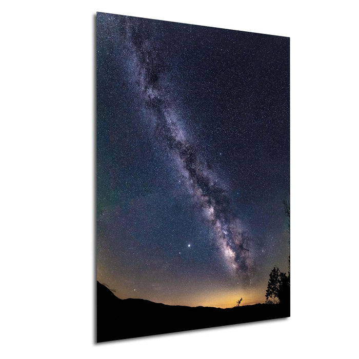 a stunning night sky with the Milky Way galaxy stretching across the frame, its bright white center contrasting against the dark blue backdrop. The silhouettes of trees can be seen in the distance, adding a sense of depth and scale to the scene.