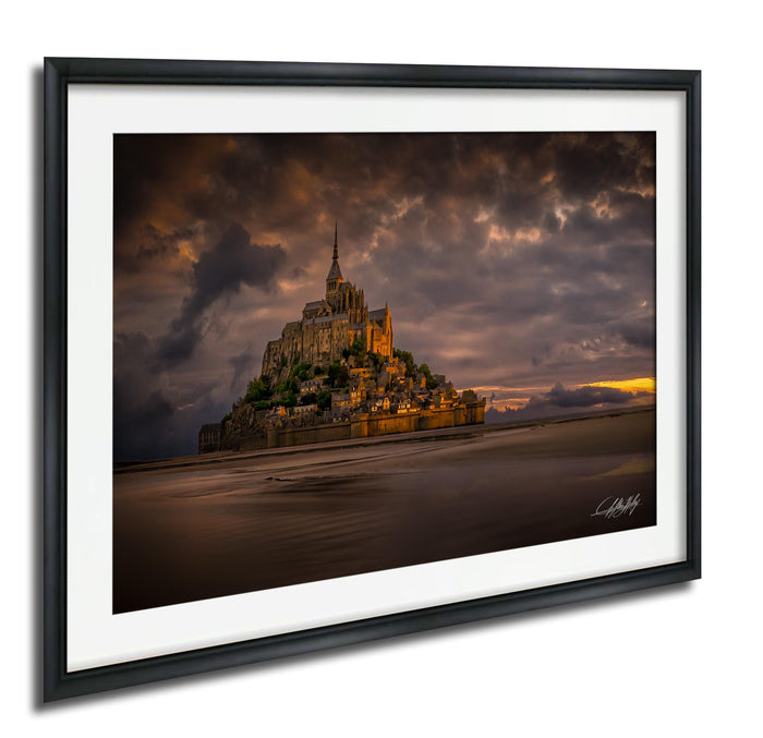 A framed photograph depicting the iconic Mont Saint-Michel, a medieval fortified island in Normandy, France, with its distinctive spire and castle-like architecture set against a dramatic sky.