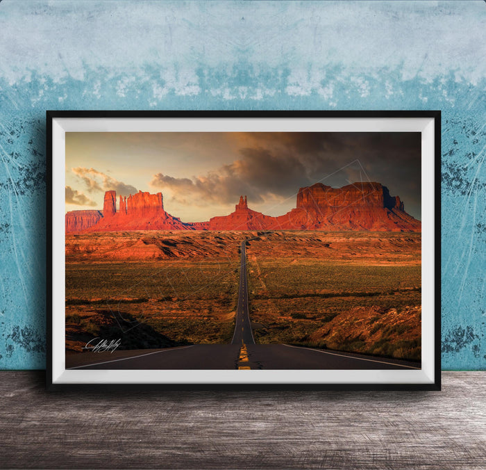 A framed photograph depicting a desert landscape with a winding road leading towards a series of red rock formations in the distance, set against a cloudy sky.