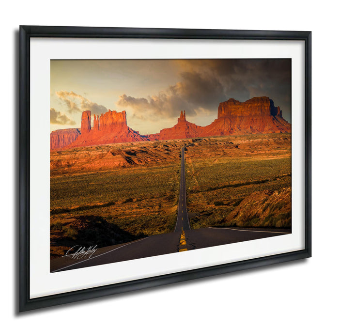 a long, straight road cutting through a vast, arid landscape with towering red rock formations in the background.
