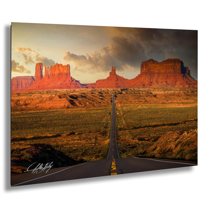 a long, straight road cutting through a vast, arid landscape with towering red rock formations in the background. The sky above is filled with clouds, creating a dramatic atmosphere. The road appears to be the main focus of the image, leading the viewer's eye towards the distant mountains.