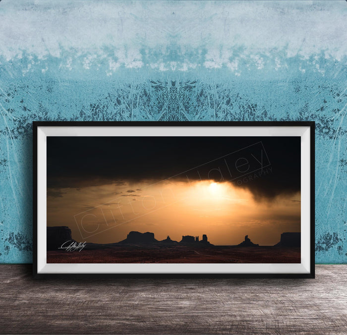 a framed photograph of a desert landscape at sunset, with silhouetted mountains in the background and a dramatic sky filled with clouds and the sun setting behind them.